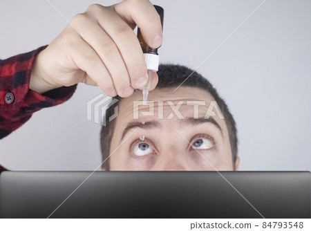 Close-up of eye drops. Artificial tear. Pain in the eyes. Sensation of grit in the eyes. A man looks out from behind a computer monitor or laptop. Drip my own eye medicine Close-up of eye drops. Artificial tear. Pain in the eyes. Sensation of grit in the eyes. A man looks out from behind a computer monitor or laptop. Drip my own eye medicine 84793548