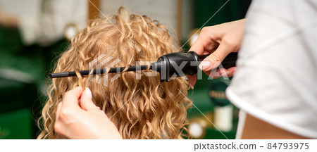 Hands of hairstylist curl wavy hair of young woman using a curling iron for hair curls in the beauty salon rear view. Hands of hairstylist curl wavy hair of young woman using a curling iron for hair curls in the beauty salon rear view. 84793975