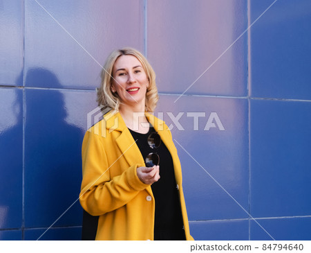 Young woman with blond hair in a bright yellow coat on a blue background on a city street. Urban style of modern woman Young woman with blond hair in a bright yellow coat on a blue background on a city street. Urban style of modern woman 84794640