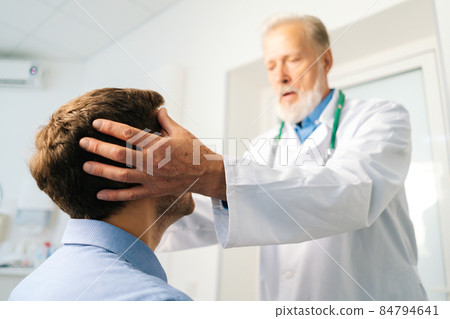 Close-up low-angle view of mature adult male doctor massaging temples to sick patient during consultation at hospital. 84794641