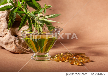 Gravy boat or jug and capsules with hemp oil, branch with leaf of cannabis on natural stone on craft paper, with copy space. Gravy boat or jug and capsules with hemp oil, branch with leaf of cannabis on natural stone on craft paper, with copy space. 84796027