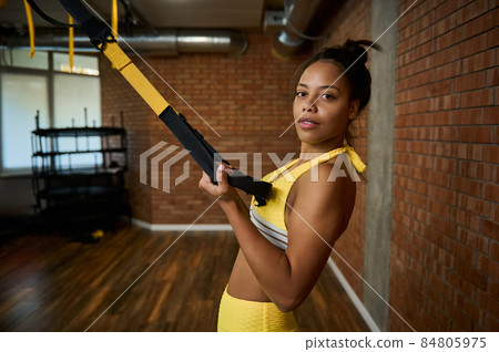 Beautiful African woman doing arm exercises with suspension straps at gym. Healthy and active lifestyle, body care, fitness and sport concept Beautiful African woman doing arm exercises with suspension straps at gym. Healthy and active lifestyle, body care, fitness and sport concept 84805975