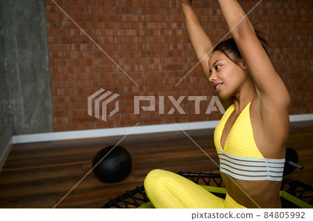 Young African woman in bright yellow tracksuit raising arms up, stretching her body, sitting in lotus position on trampoline and looking at her mirror reflection while working out in fitness studio Young African woman in bright yellow tracksuit raising arms up, stretching her body, sitting in lotus position on trampoline and looking at her mirror reflection while working out in fitness studio 84805992