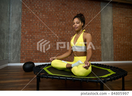 Beautiful young African woman in bright yellow tracksuit sitting on a trampoline in lotus pose and smiles looking at camera. Fitness, sport, meditation, yoga, active lifestyle concept 84805997