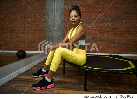 African woman with slim body and flat belly in bright yellow tracksuit sitting on a trampoline in lotus pose and smiles looking at camera. Fitness, sport, meditation, yoga, active lifestyle concept 84805998