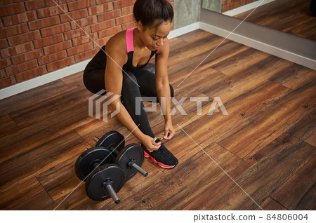 Overhead view of a young African American female athlete, attractive woman with perfect muscular body tying shoelaces on her sneakers, next to dumbbells lying down on the floor of a gym Overhead view of a young African American female athlete, attractive woman with perfect muscular body tying shoelaces on her sneakers, next to dumbbells lying down on the floor of a gym 84806004