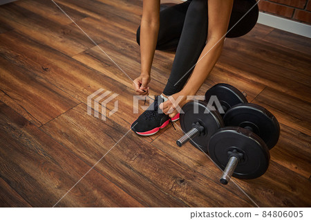 Close-up of a female athlete,sporty woman with perfect muscular body tying shoelaces on her sneakers, next to dumbbells lying down on the floor of a gym 84806005
