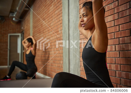 Beautiful athletic slim body woman in stylish tight-fitting tracksuit, ties her ponytail before starting a workout, sitting on a sports ball against a large mirror with her blurred reflection Beautiful athletic slim body woman in stylish tight-fitting tracksuit, ties her ponytail before starting a workout, sitting on a sports ball against a large mirror with her blurred reflection 84806006