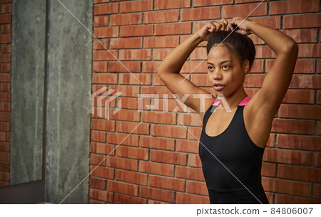 Beautiful athletic African American woman in stylish tight-fitting tracksuit, ties her ponytail before starting a workout, sitting on a sports ball against a large mirror with her blurred reflection Beautiful athletic African American woman in stylish tight-fitting tracksuit, ties her ponytail before starting a workout, sitting on a sports ball against a large mirror with her blurred reflection 84806007