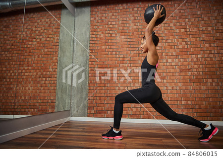 Young African woman, athlete with a beautiful muscular body doing exercises on the buttocks - lunges, holding a medical gymnastic ball on outstretched arms over her head while training in the gym 84806015