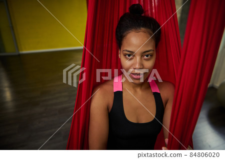 Close-up portrait of African woman with slim body in a black activewear practicing aerial fly yoga on a red hammock Close-up portrait of African woman with slim body in a black activewear practicing aerial fly yoga on a red hammock 84806020