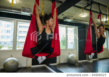 Beautiful serene African American young woman practices aerial antigravity yoga, looks at her mirror reflection in yoga class Beautiful serene African American young woman practices aerial antigravity yoga, looks at her mirror reflection in yoga class 84806025