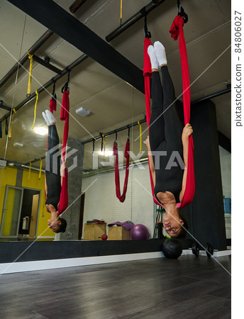 Charming sportive young African American fit woman practicing aerial flying yoga, doing stretching exercises hanging upside down on red hammock in yoga studio Charming sportive young African American fit woman practicing aerial flying yoga, doing stretching exercises hanging upside down on red hammock in yoga studio 84806027
