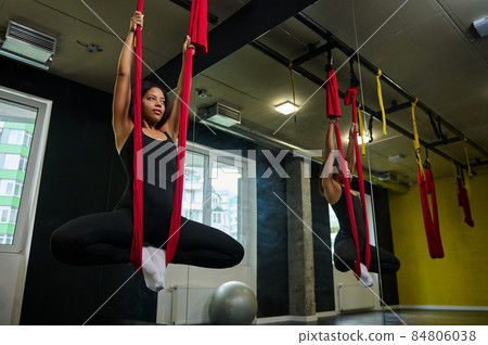 Young African fitness woman in sportswear practicing fly yoga in studio, stretching body with red hammock or swing. Aerial aero fly fitness. Body care and mindfulness 84806038