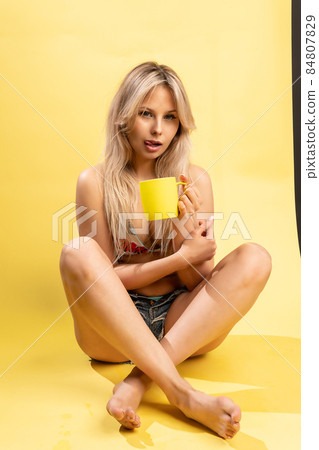 A girl sitting on the floor with her feet cross-legged keeps her head on a beautiful yellow background with a cup in hand yellow or pink and red playful cheerful beauty, young beautiful body posing A girl sitting on the floor with her feet cross-legged keeps her head on a beautiful yellow background with a cup in hand yellow or pink and red playful cheerful beauty, young beautiful body posing 84807829