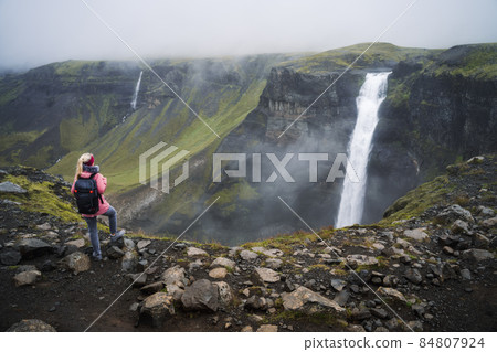 Woman with backpack and lilac jacket enjoying Haifoss waterfall of Iceland Highlands in Thjorsardalur Valley Woman with backpack and lilac jacket enjoying Haifoss waterfall of Iceland Highlands in Thjorsardalur Valley 84807924