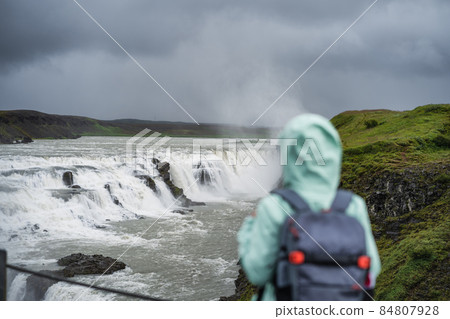 Woman with backpack and green jacket standing in front of Gullfoss powerful famous waterfall in Iceland. Tourist attraction who follow Golden Circle route of Iceland 84807928