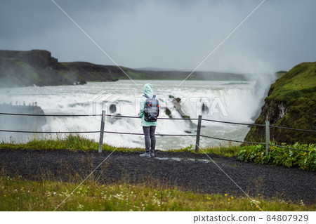 Woman with backpack and green jacket visit Gullfoss powerful famous waterfall in Iceland. Tourist attraction who follow Golden Circle route of Iceland 84807929