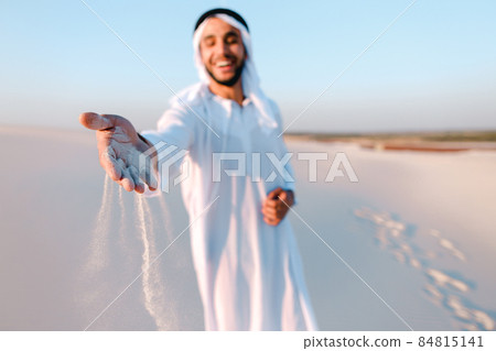 Close-up shot of portrait and hands of young Arab guy in sandy desert on clear summer day in open air. Close-up shot of portrait and hands of young Arab guy in sandy desert on clear summer day in open air. 84815141