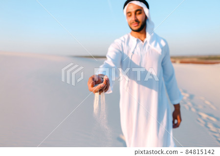 Close-up shot of portrait and hands of young Arab guy in sandy desert on clear summer day in open air. 84815142