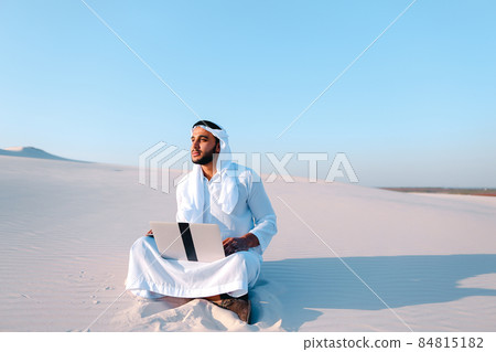Emirati guy professional designer with computer projects project, sitting on sand in desert on summer evening. Emirati guy professional designer with computer projects project, sitting on sand in desert on summer evening. 84815182