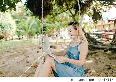 Young happy girl using laptop and riding on swing on sand. Young happy girl using laptop and riding on swing on sand. 84816117