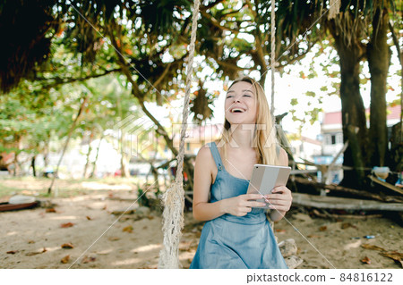 Young smiling girl using tablet and riding swing on sand, wearing jeans sundress. 84816122