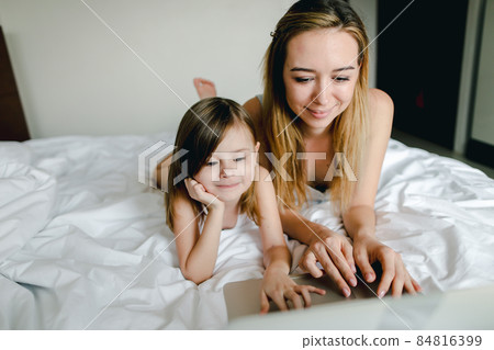 Young successful woman using laptop with little daughter and lying in bed. 84816399