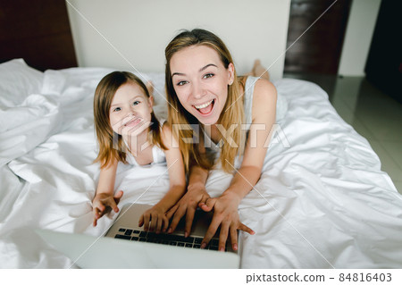 Happy blonde female person using laptop with little daughter and lying in bed. 84816403