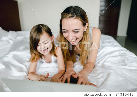 Happy female person using laptop with little daughter and lying in bed. 84816415