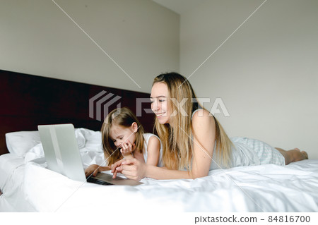 Young happy mother lying in bed with little daughter and using laptop. 84816700