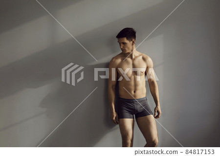 Sexy portrait of muscular shirtless male model in underwear posing in sensual pose against gray wall background. Caucasian brunette man in black cotton underpants looks away while standing in sunlight Sexy portrait of muscular shirtless male model in underwear posing in sensual pose against gray wall background. Caucasian brunette man in black cotton underpants looks away while standing in sunlight 84817153