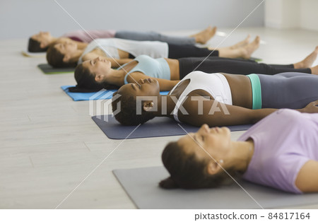 Women on group practicing yoga. Multiracial young women lie on floor and practice deep breathing exercises in yoga class. Shavasana or corps pose is the end of a class or practice. Selective focus. Women on group practicing yoga. Multiracial young women lie on floor and practice deep breathing exercises in yoga class. Shavasana or corps pose is the end of a class or practice. Selective focus. 84817164