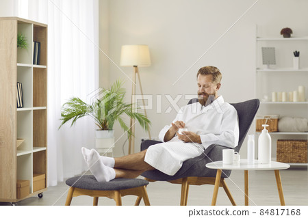 Male beauty. Cheerful bearded man enjoys cosmetic procedures at home by applying moisturizing mask on his face. Funny man in bathrobe and white socks sits on chair and tries out various cosmetics. Male beauty. Cheerful bearded man enjoys cosmetic procedures at home by applying moisturizing mask on his face. Funny man in bathrobe and white socks sits on chair and tries out various cosmetics. 84817168