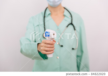 Temperature measurement gun in doctor hands. Close-up shot of doctor wearing protective surgical mask ready to use infrared isometric thermometer gun to check body temperature for virus symptoms. 84822020