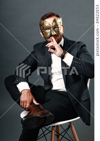 Man in business suit sits on chair wearing carnival mask on his face. Studio portrait on gray 84822653