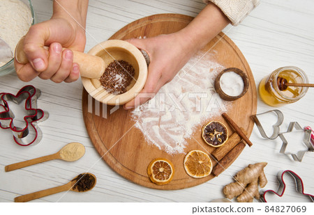 Woman's hands grinding ingredients in a marble mortar standing on a wooden board with sprinkled flour and dried orange slices, cinnamon sticks lying on it, cutting molds on a wooden table 84827069