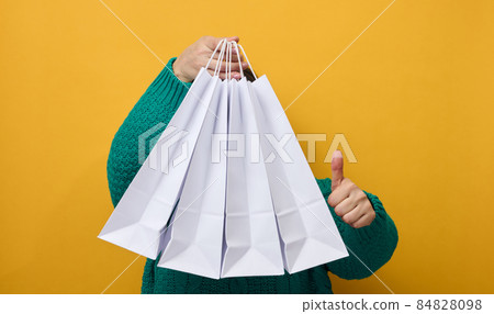 a woman in a sweater holds white paper bags on a yellow background and shows a like gesture with her hand. Purchases. Seasonal sale 84828098