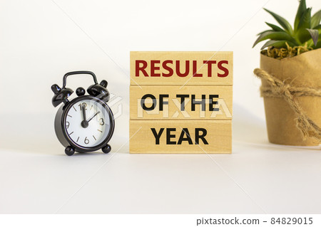 Results of the year symbol. Wooden blocks with words 'Results of the year'. White table. Black alarm clock and house plant. Beautiful white background. Copy space. Business and results of the year Results of the year symbol. Wooden blocks with words 'Results of the year'. White table. Black alarm clock and house plant. Beautiful white background. Copy space. Business and results of the year 84829015