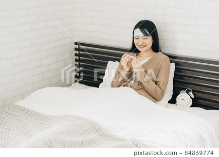 A smiling woman measuring her body temperature using a thermometer in her bedroom (cooling sheet) A smiling woman measuring her body temperature using a thermometer in her bedroom (cooling sheet) 84839774