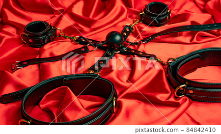 Adult sex games. BDSM items. Leather straps handcuffs and gag ball on a red satin sheet. Adult sex games. BDSM items. Leather straps handcuffs and gag ball on a red satin sheet. 84842410