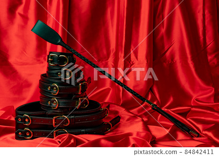 Adult sex games. BDSM items. Leather straps handcuffs, belt and whip on a red satin sheet. Adult sex games. BDSM items. Leather straps handcuffs, belt and whip on a red satin sheet. 84842411