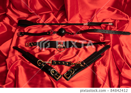 Adult sex games. BDSM items. Leather straps handcuffs, collar, gag ball and whip on a red sheet. Adult sex games. BDSM items. Leather straps handcuffs, collar, gag ball and whip on a red sheet. 84842412