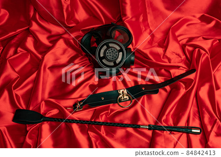 Adult sex games. BDSM items. Gas mask, collar and whip on a red satin sheet. Adult sex games. BDSM items. Gas mask, collar and whip on a red satin sheet. 84842413