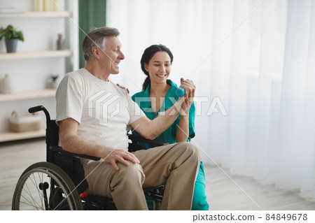 Young female physiotherapist helping senior man in wheelchair to work out with dumbbells, to recover from injury 84849678