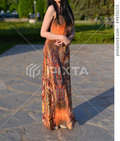 beautiful brunette model with long hair is having a photo shooting in the park. the woman is wearing an orange long summer dress beautiful brunette model with long hair is having a photo shooting in the park. the woman is wearing an orange long summer dress 84850215