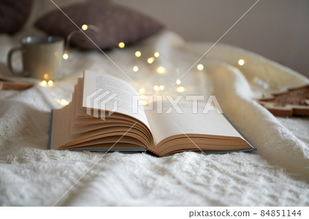 Opened book on a knited beige plaid, christmas garland lights and mug, cozy winter day at home 84851144