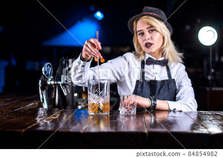 Focused girl bartending pouring fresh alcoholic drink into the glasses behind the bar Focused girl bartending pouring fresh alcoholic drink into the glasses behind the bar 84854982