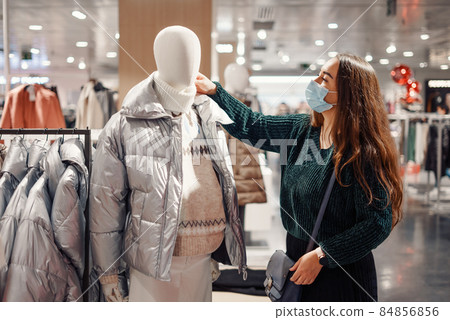 Young lady in face mask chooses warm clothes in department store, standing near mannequin dressed in puffy winter jacket and sweater, buying new seasonal outfit. Shopping during covid19 pandemic Young lady in face mask chooses warm clothes in department store, standing near mannequin dressed in puffy winter jacket and sweater, buying new seasonal outfit. Shopping during covid19 pandemic 84856856