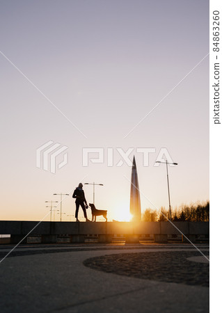 woman walking her dog at sunset, beautiful sunset against the background of modern buildings woman walking her dog at sunset, beautiful sunset against the background of modern buildings 84863260